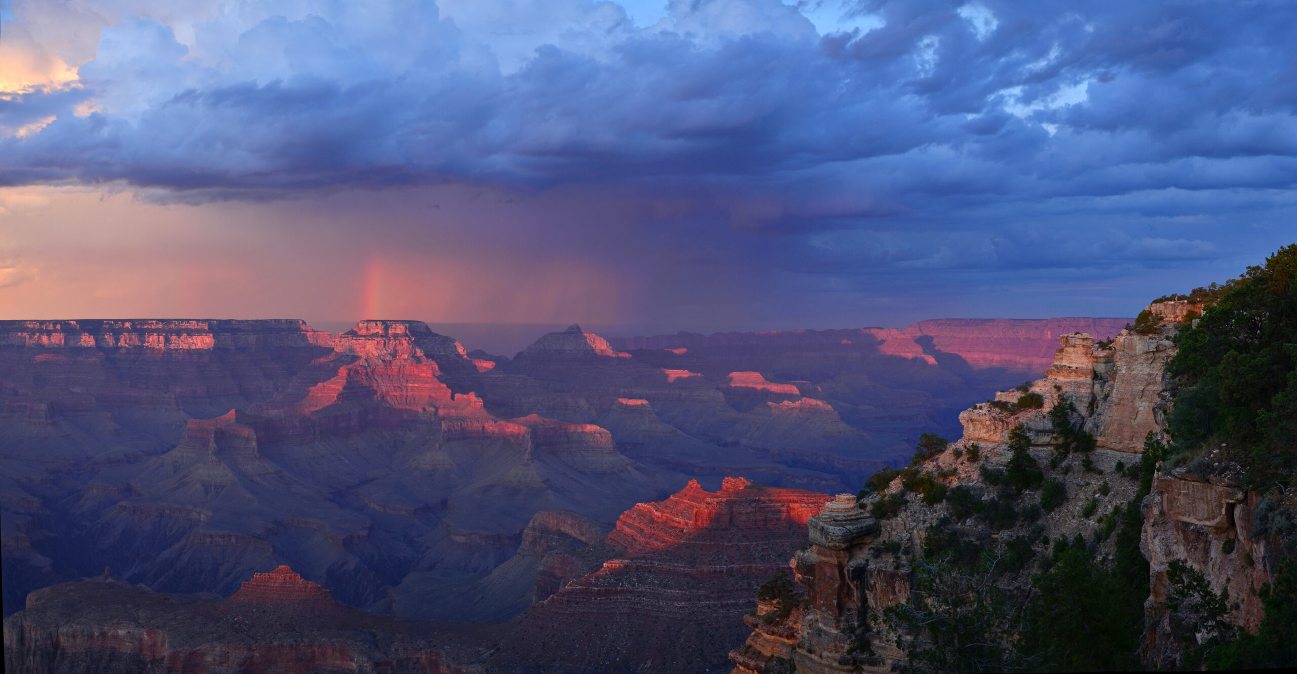 Moody sunset photo of the Grand Canyon with low-hanging clouds, rain and portion of a rainbow in the distance, with low-angled sun illuminating buttes and cliffs.